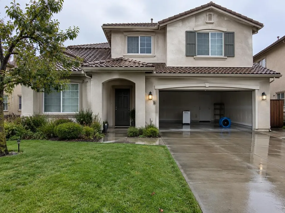 Generated illustration of a two-story stucco house after rain with drying equipment in the garage